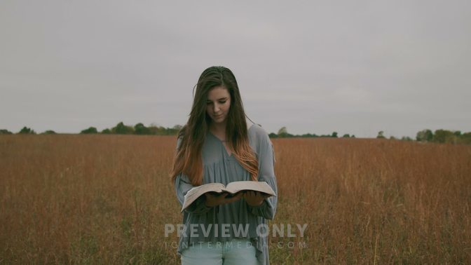 A Woman Walking Through A Field Of Tall Grasses Reading A Bible Stock